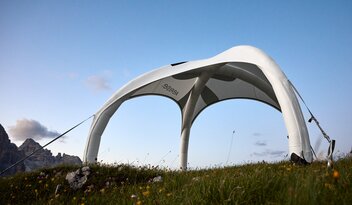 Ein aufblasbares Zelt in der Farbe Weiß, das auf einer Bergwiese steht und aus Froschperspektive fotografiert wurde.