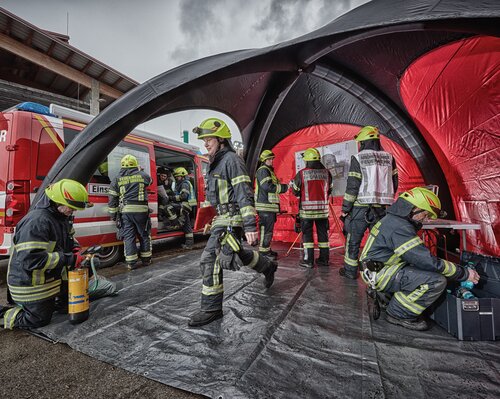 Schwarz-rotes Luftzelt, unter dem mehrere Personen der Feuerwehr arbeiten. Links daneben ein Einsatzwagen.