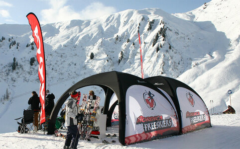 Zwei schwarze aufblasbare Eventzelte mit bedruckten weißen Seitenwände, aufgebaut auf beschneiter Skipiste