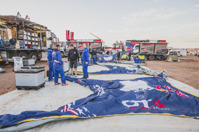 Crew in blauer Kleidung entfaltet ein aufblasbares Zelt vor Servicefahrzeugen im Rally-Lager.